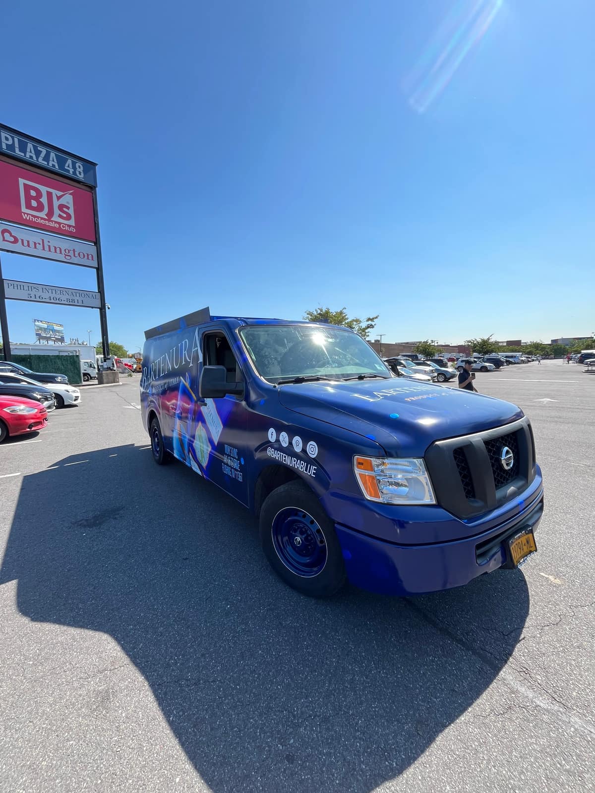 Blue Nissan NV cargo van in a full printed wrap for Bartenura, parked in daylight in a retail plaza lot with big-box store signage behind it