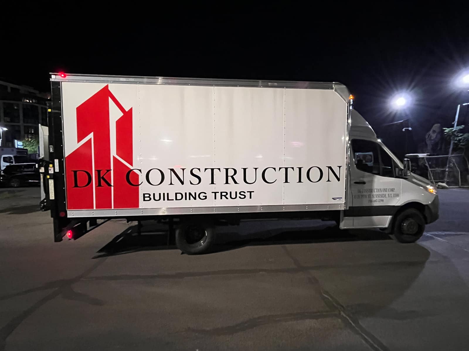 White Mercedes Sprinter box truck lettered for DK Construction of Sunnyside, Queens, with a red printed logo on the box, parked in a lot at night